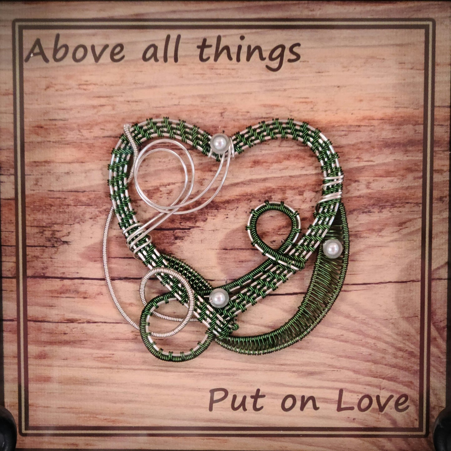 Green & Silver woven wire heart in light wood shadow box and Background print of wood planks is printed on canvas medium and reads "Above all things, Put on Love."