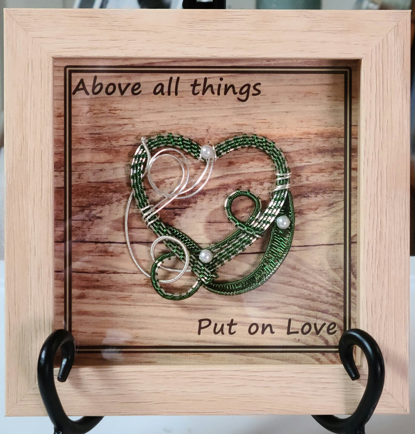 Green & Silver woven wire heart in light wood shadow box and Background print of wood planks is printed on canvas medium and reads "Above all things, Put on Love."