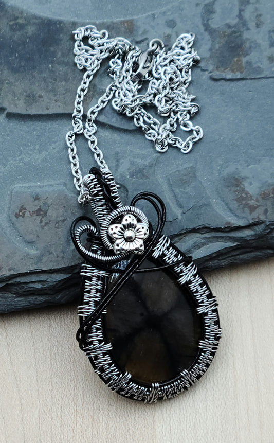 This chiastolite stone is a soft brown with an interesting, off-center drown/black cross. It's really very interesting. I've framed it with tarnish resistant silver plated copper wire in an color called titanium woven over enameled black base wires. Then I have it a small pewter flower accent.