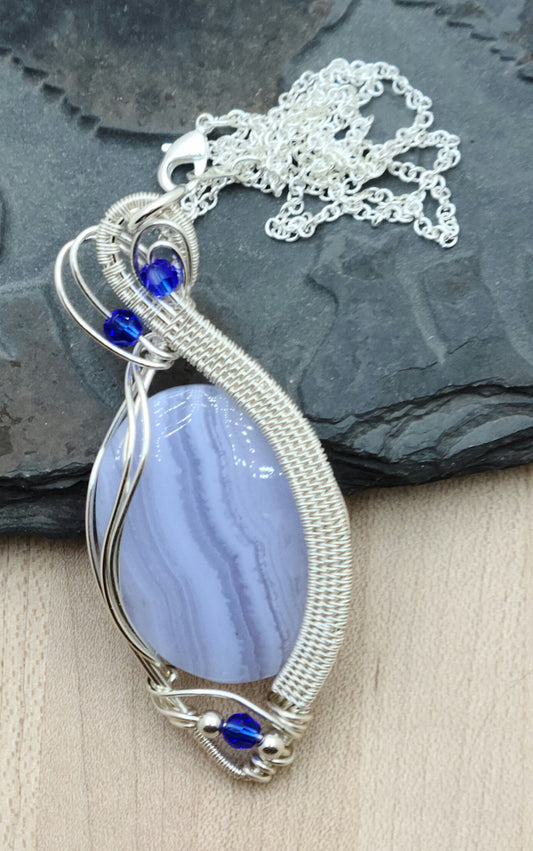 Enjoy the calming effect of this lovely blue lace agate stone pendant, with its frame woven from tarnish resistant silver plated copper & adorned with bright blue crystals.