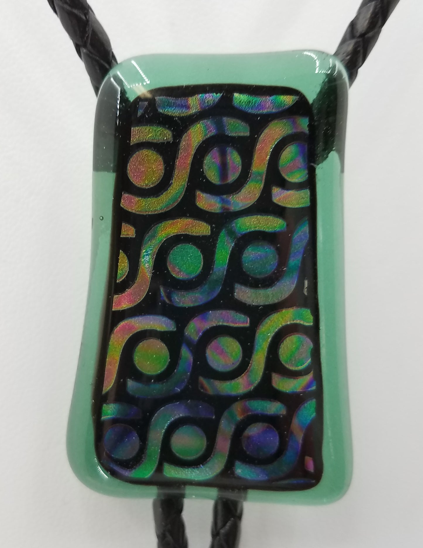 bolo tie of Geometric gold with black, green, & purple dichroic glass backed by translucent green glass with black leather
