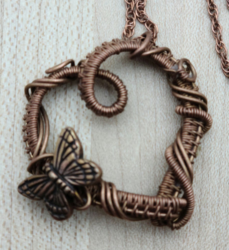 Expertly handcrafted with antiqued copper, this woven wire heart pendant is adorned with a delicate butterfly bead, making it a stunning statement piece. Perfect for any occasion, its unique design is sure to garner attention.