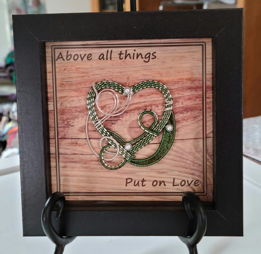 Green & Silver woven wire heart in black shadow box and Background print of wood planks is printed on canvas medium and reads "Above all things, Put on Love."