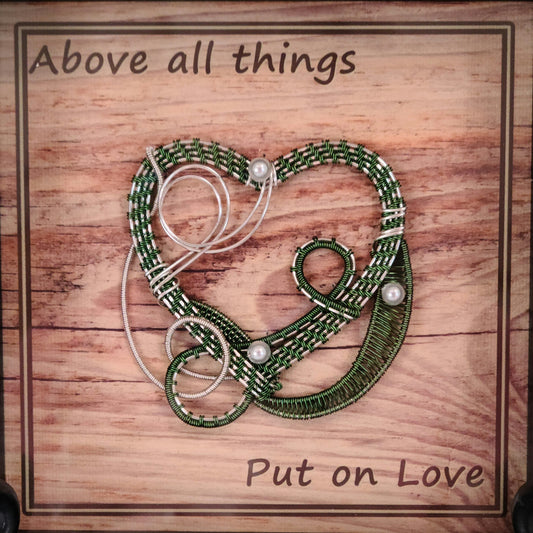 Green & Silver woven wire heart in black shadow box and Background print of wood planks is printed on canvas medium and reads "Above all things, Put on Love."