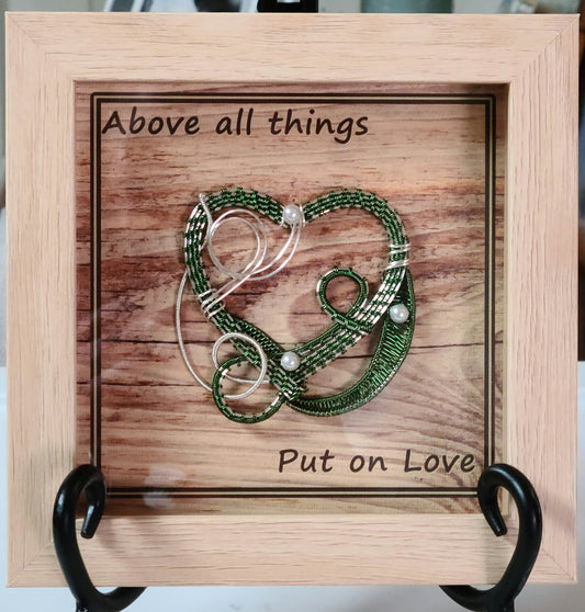 Green & Silver woven wire heart in light wood shadow box and Background print of wood planks is printed on canvas medium and reads "Above all things, Put on Love."