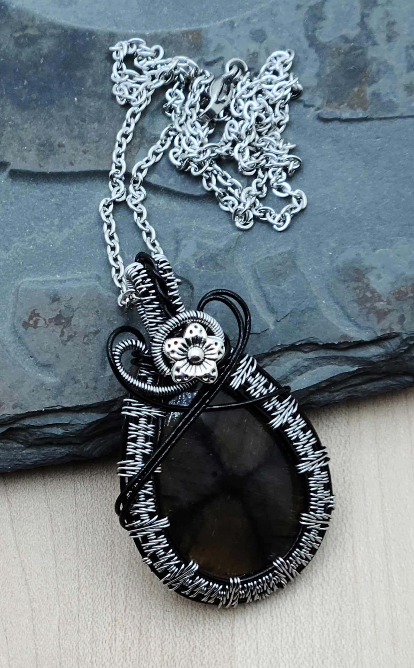 This chiastolite stone is a soft brown with an interesting, off-center drown/black cross. It's really very interesting. I've framed it with tarnish resistant silver plated copper wire in an color called titanium woven over enameled black base wires. Then I have it a small pewter flower accent.