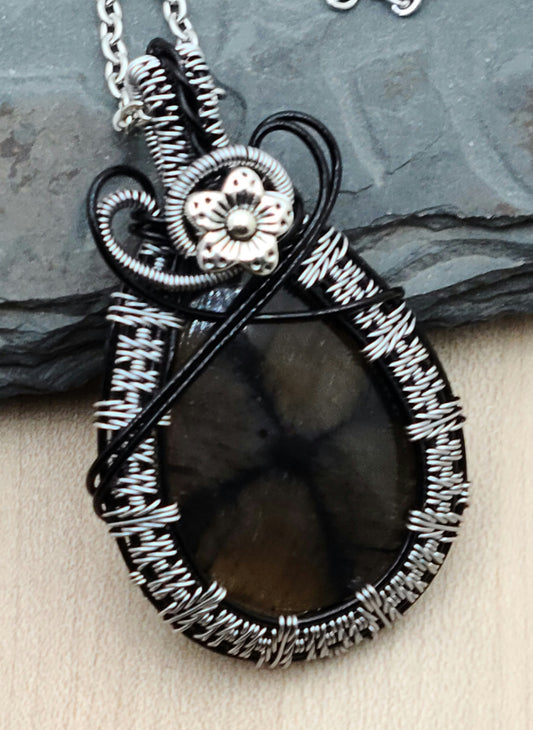 This chiastolite stone is a soft brown with an interesting, off-center drown/black cross. It's really very interesting. I've framed it with tarnish resistant silver plated copper wire in an color called titanium woven over enameled black base wires. Then I have it a small pewter flower accent.