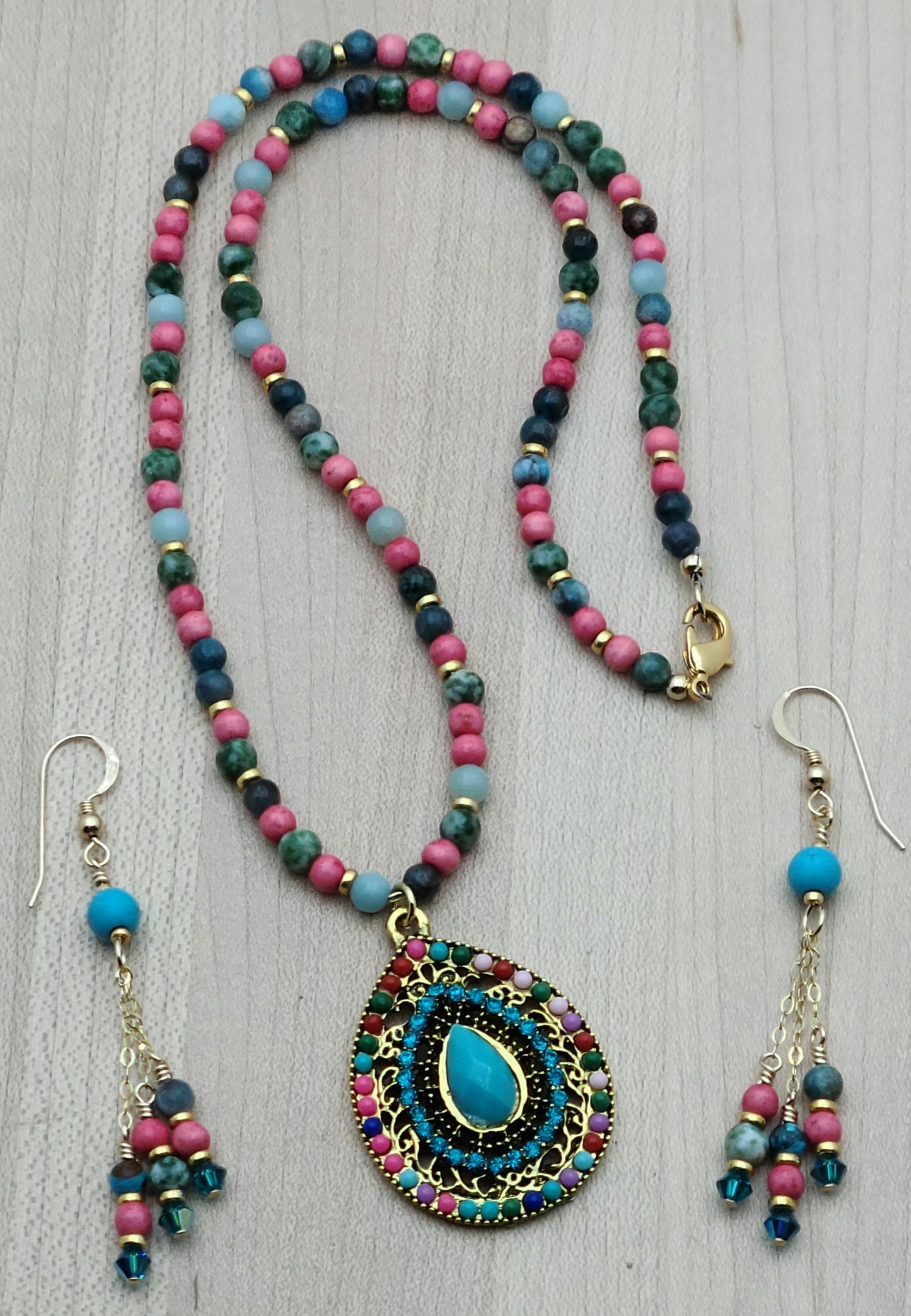 Fun and vibrant, this pendant necklace and earring set captures attention with its bold design and bright, happy colors of turquoise, bright pink, green, & gold.