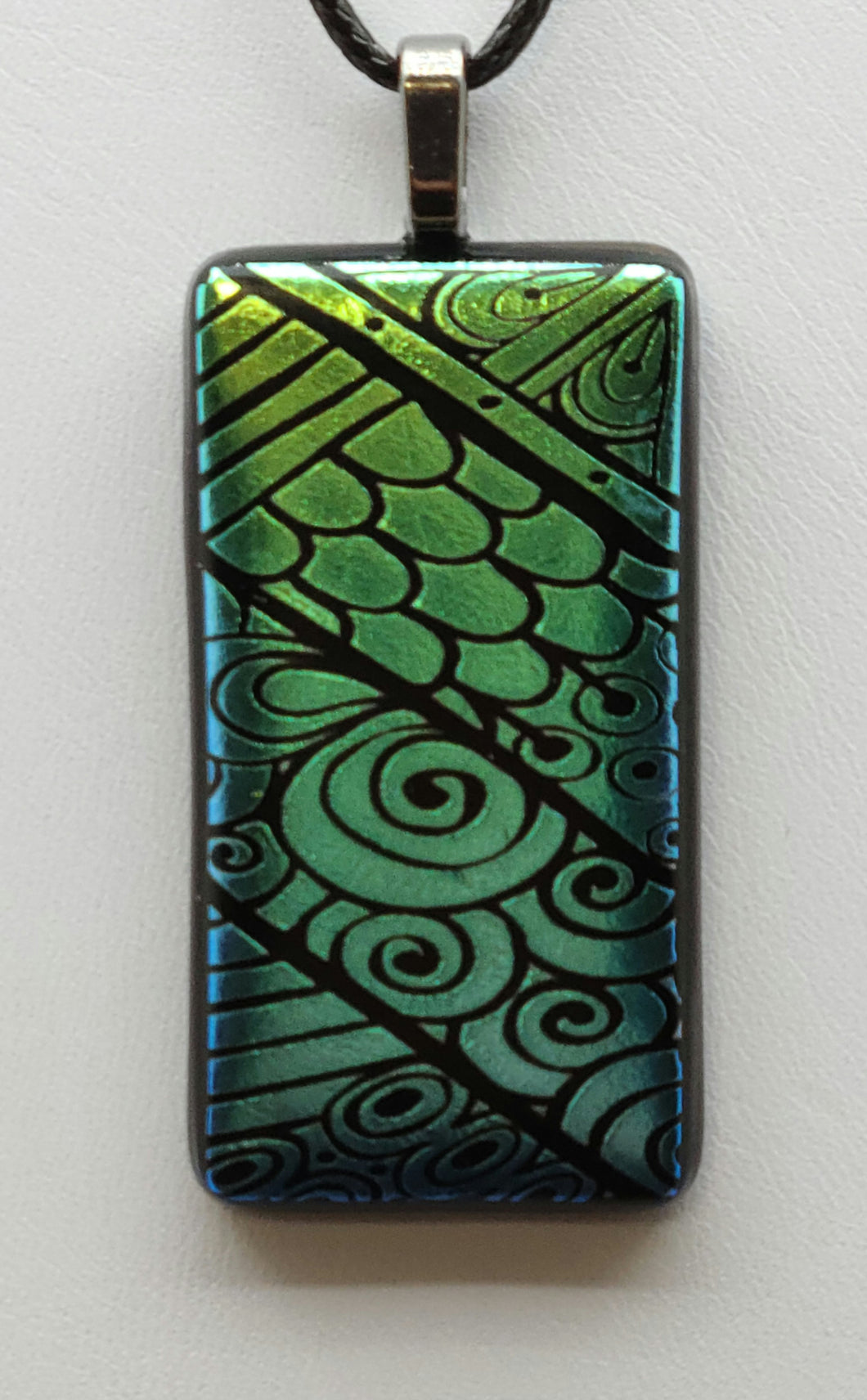 This fused glass pendant is a whimsical combination of scales, lines, and curly-q's etched into dichroic glass that magically shifts from teal to green.